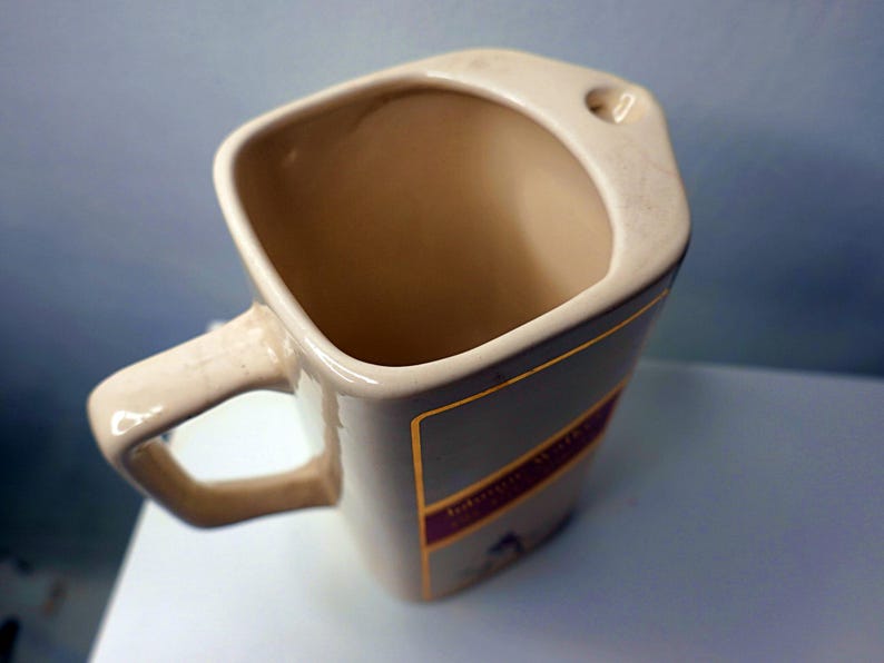 May include: Tan ceramic mug with a square shape and a handle. The mug has a label with gold and purple accents. The interior is a light tan color. The mug is sitting on a white surface.