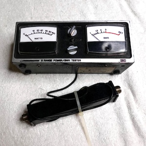 May include: A black and silver 8-range power/SWR tester with two analog meters. The left meter reads "WATTS" and the right meter reads "SWR". A black cable and connector are attached. The text "8 RANGE POWER/SWR TESTER" is visible.