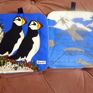 Puede incluir: Dos agarraderas de olla de tela azul y blanca. Una presenta dos frailecillos en una roca con hierba verde y un fondo blanco. La otra presenta narvales nadando en agua azul con un iceberg blanco en el fondo. Ambas agarraderas de olla tienen un borde azul y una etiqueta blanca con la palabra "Freeman" y un número.