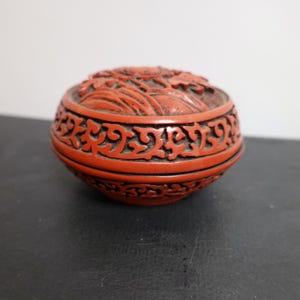 May include: A round, red decorative box with intricate carved floral and scrollwork patterns. The box has a lid with a raised design and a detailed band around the middle. The overall design is symmetrical and ornate.