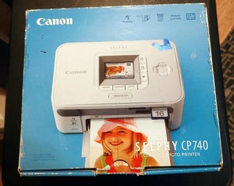 May include: A white Canon Selphy CP740 compact photo printer is displayed in its original box. The box is blue with the Canon logo and printer features. A photo is being printed, showing a girl wearing a hat.