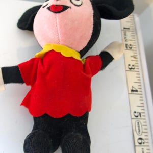 K&ouml;nnte beinhalten: Ein Vintage-Pl&uuml;schtier mit einer Zeichentrickfigur mit rosa Gesicht, schwarzen Ohren und einer gelben Schleife. Die Figur tr&auml;gt ein rotes Hemd mit gewelltem Saum und eine schwarze Hose. Das Spielzeug ist etwa 15 cm gro&szlig;.