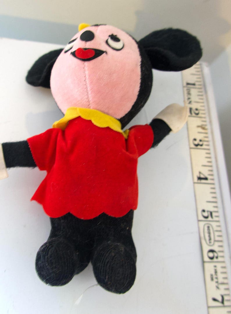 K&ouml;nnte beinhalten: Ein Vintage-Pl&uuml;schtier mit einer Zeichentrickfigur mit rosa Gesicht, schwarzen Ohren und einer gelben Schleife. Die Figur tr&auml;gt ein rotes Hemd mit gewelltem Saum und eine schwarze Hose. Das Spielzeug ist etwa 15 cm gro&szlig;.