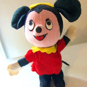 K&ouml;nnte beinhalten: Eine Vintage-Pl&uuml;schmaus mit gro&szlig;en schwarzen Ohren, einem rosa Gesicht und einem roten Kleid. Die Maus hat einen gelben Kragen und einen schwarzen Schwanz. Ihre Arme sind in einer winkenden Geste erhoben, mit wei&szlig;en Handschuhen. Ein klassisches Kinderspielzeug.