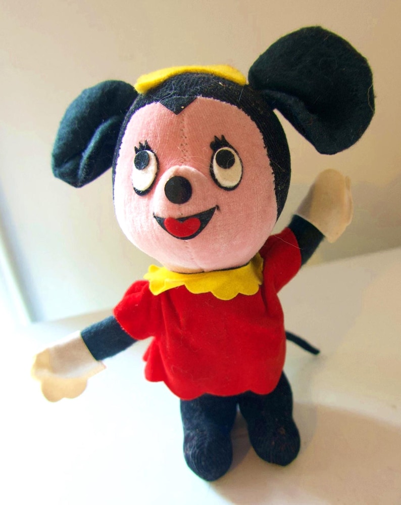 K&ouml;nnte beinhalten: Eine Vintage-Pl&uuml;schmaus mit gro&szlig;en schwarzen Ohren, einem rosa Gesicht und einem roten Kleid. Die Maus hat einen gelben Kragen und einen schwarzen Schwanz. Ihre Arme sind in einer winkenden Geste erhoben, mit wei&szlig;en Handschuhen. Ein klassisches Kinderspielzeug.