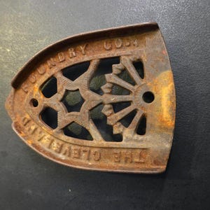 Op de afbeelding: Een roestige, antieke strijkijzerbasis met een opengewerkt ontwerp. De tekst "THE CLEVELAND LAUNDRY CO." is zichtbaar. De strijkijzerbasis heeft een donkerbruine kleur, met een ster- en wielpatroon.