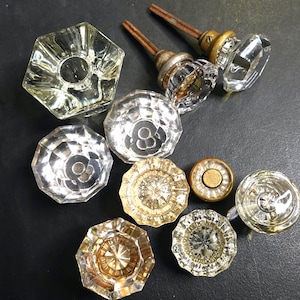 Op de afbeelding: Een verzameling vintage glazen deurknoppen in verschillende vormen en maten. De knoppen zijn helder, facetgeslepen en sommige hebben messing of metalen stelen. De opstelling omvat ronde, achthoekige en andere geometrische ontwerpen, die een verscheidenheid aan antieke hardware laten zien.