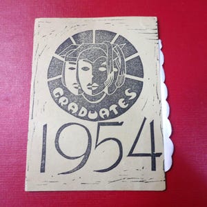 May include: A vintage yearbook cover featuring a black and white graphic of three faces within a circle, the word "GRADUATES" and the year "1954". The cover rests on a red surface and has a scalloped edge.
