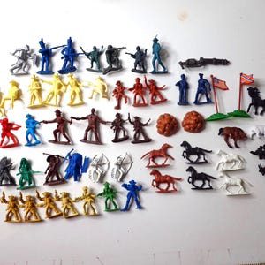 Könnte beinhalten: Eine Sammlung von Vintage-Spielzeugsoldaten, Pferden und anderen Figuren aus Kunststoff in verschiedenen Farben. Die Figuren umfassen Soldaten, Cowboys und Indianer sowie eine Kanone und Flaggen. Das Spielzeug ist auf einer weißen Oberfläche angeordnet.