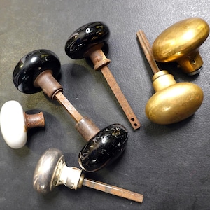 May include: A collection of vintage door knobs in various styles. Includes black, white, and brass knobs with aged, rusty metal stems. The knobs are oval and round, showcasing a range of finishes and designs. Ideal for restoration projects.