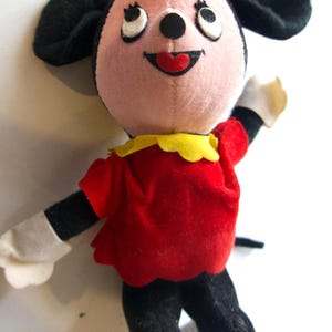 K&ouml;nnte beinhalten: Eine Vintage-Pl&uuml;schpuppe mit einer Zeichentrickfigur mit rosa Gesicht, schwarzen Ohren und einer gelben Schleife. Die Puppe tr&auml;gt ein rotes Kleid mit gelbem Kragen, schwarze Beine und wei&szlig;e Handschuhe. Die Augen der Puppe blicken nach oben.