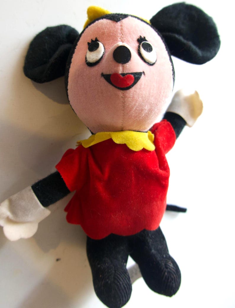K&ouml;nnte beinhalten: Eine Vintage-Pl&uuml;schpuppe mit einer Zeichentrickfigur mit rosa Gesicht, schwarzen Ohren und einer gelben Schleife. Die Puppe tr&auml;gt ein rotes Kleid mit gelbem Kragen, schwarze Beine und wei&szlig;e Handschuhe. Die Augen der Puppe blicken nach oben.