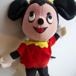 K&ouml;nnte beinhalten: Ein Vintage-Pl&uuml;schtier mit einem l&auml;chelnden Cartoon-Charakter mit schwarzen Ohren, einem rosa Gesicht und einem roten Kleid. Das Spielzeug hat wei&szlig;e Handschuhe, schwarze Beine sowie einen gelben Kragen und Hut. Die Augen der Figur sind gro&szlig; und wei&szlig; mit schwarzen Pupillen.