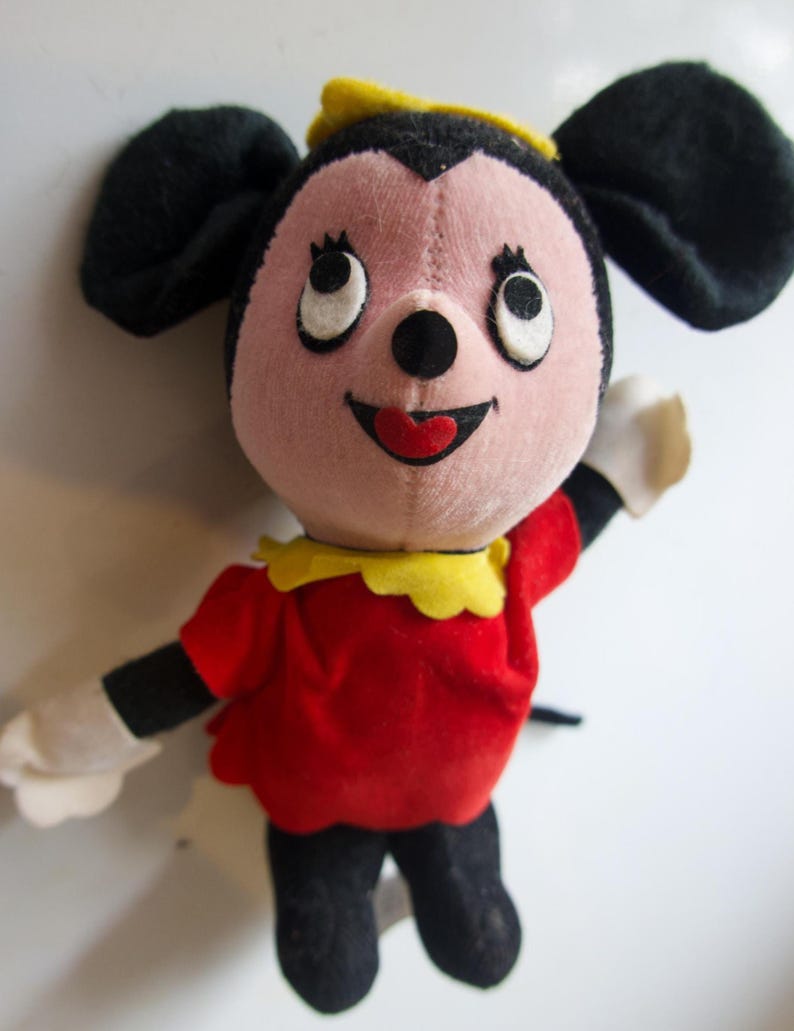 K&ouml;nnte beinhalten: Ein Vintage-Pl&uuml;schtier mit einem l&auml;chelnden Cartoon-Charakter mit schwarzen Ohren, einem rosa Gesicht und einem roten Kleid. Das Spielzeug hat wei&szlig;e Handschuhe, schwarze Beine sowie einen gelben Kragen und Hut. Die Augen der Figur sind gro&szlig; und wei&szlig; mit schwarzen Pupillen.