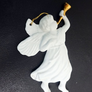 Könnte beinhalten: Weißer Porzellanengel-Anhänger mit goldenen Flügeln und einer goldenen Trompete. Der Engel spielt Trompete und trägt ein weißes Kleid.