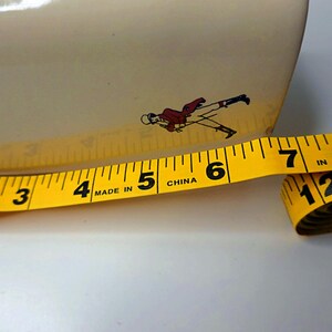 May include: A yellow measuring tape with black markings, showing inches, is partially unrolled. The tape is next to a cream-colored object with a small illustration of a person in a red coat. The tape reads "MADE IN CHINA".