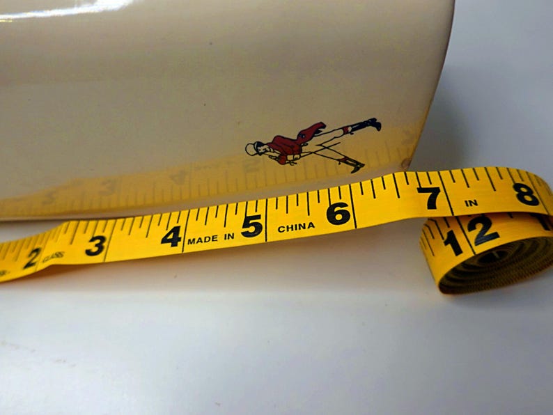 May include: A yellow measuring tape with black markings, showing inches, is partially unrolled. The tape is next to a cream-colored object with a small illustration of a person in a red coat. The tape reads "MADE IN CHINA".