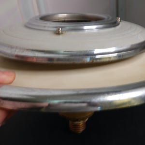 May include: A vintage, two-tiered lamp shade with a cream-colored base and a smaller white top. Both tiers are trimmed with a silver metal band. The lamp shade is held by a hand, showcasing its size and design.