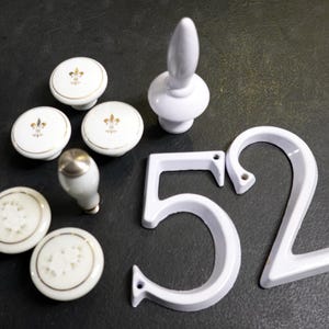 May include: White ceramic cabinet knobs with gold fleur-de-lis designs, a white door handle, and white metal house numbers 5 and 2. The knobs have a gold trim. The items are arranged on a dark surface.