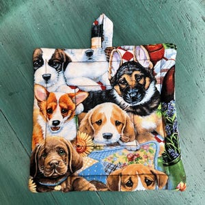 May include: A square, quilted pot holder featuring a collage of adorable puppies. The puppies are various breeds and colors, including brown, black, and white. The pot holder has a loop for hanging and is set against a green background.