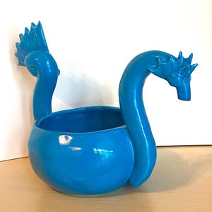 May include: A bright blue ceramic bowl featuring two sculpted necks extending upward from the sides. One neck terminates in a fin-like crest, while the other features a dragon-like head with a pointed snout and a spiky crown-like ridge on top.