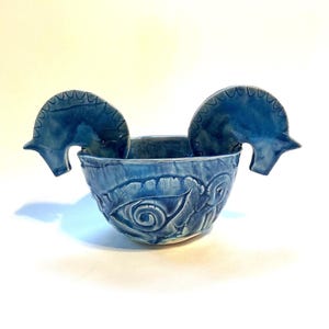 May include: A blue ceramic bowl featuring a glossy finish and two protruding handles shaped like stylized horse heads. The exterior of the bowl is decorated with a relief pattern of an eye, and the overall surface displays variations in blue tones.