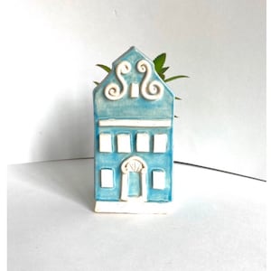 Puede incluir: Una pequeña casa de cerámica azul con bordes blancos y una puerta blanca. La casa tiene un diseño decorativo en la parte superior y es una maceta.