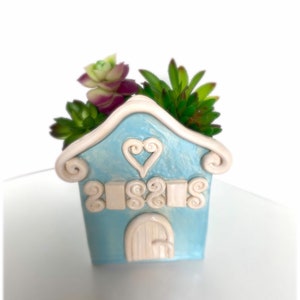 May include: A light blue ceramic house-shaped planter with white details, including a heart and a door. The planter is filled with green and purple succulents.