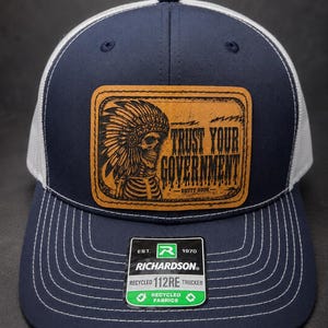 May include: A navy blue and white trucker hat featuring a rectangular tan leather patch with a black engraved graphic of a skeleton wearing a headdress. The text reads TRUST YOUR GOVERNMENT and RUSTY HOOK. A black Richardson 112RE sticker is attached.