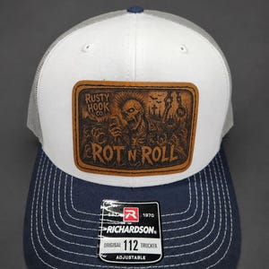 May include: A white and navy blue trucker hat featuring a rectangular brown leather patch on the front. The patch displays a graphic of a skeleton with a mohawk holding a microphone, with the text RUSTY HOOK CO. and ROT N ROLL printed on it.