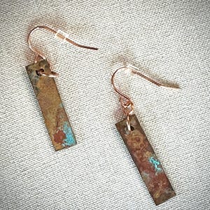 May include: A pair of dangle earrings featuring rectangular metal pendants with a weathered, oxidized finish in shades of brown, copper, and teal. The pendants hang from thin, curved copper-toned ear wires with small clear plastic backings.