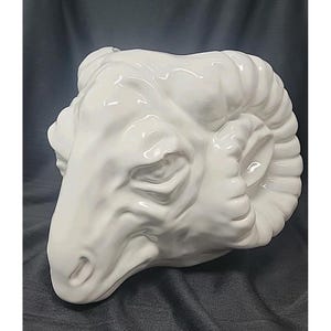May include: A white ceramic ram head sculpture. The sculpture features detailed facial features and large, curled horns. The ram's head is positioned at a slight angle, showcasing its profile. The sculpture is a decorative item.