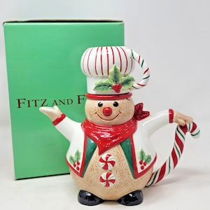 May include: A snowman-shaped teapot with a chef's hat and candy cane handle. The teapot is beige with a red scarf and green accents. The box behind the teapot has the text "FITZ AND F".