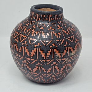 Puede incluir: Un jarrón redondo de cerámica negra con cuello estrecho y cuerpo ancho. El jarrón está decorado con un patrón geométrico repetitivo en color naranja rojizo. El interior del jarrón es de color marrón claro.