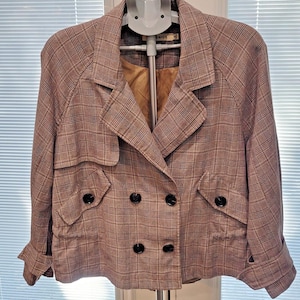 以下が含まれることがあります： モーヴ、ブラウン、クリーム色のチェック柄のクロップドダブルブレストブレザー。 ジャケットは黒いボタン、ノッチカラー、2つのフロントポケットが特徴です。 袖口は折り返されています。