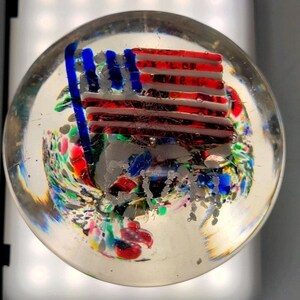 May include: A clear glass paperweight with a miniature American flag design. The flag features red and white stripes and blue stars. The base of the paperweight has colorful abstract swirls of red, green, blue, and white.