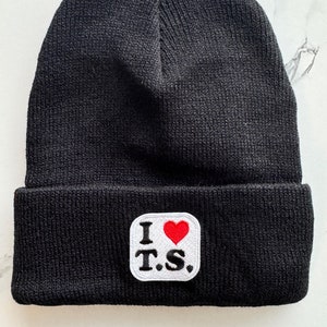 May include: Black knitted beanie with a white patch that says "I ♥ T.S." in black with a red heart.
