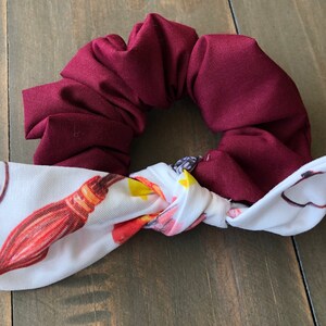 Puede incluir: Un scrunchie burdeos con un lazo blanco. El lazo tiene un estampado de una escoba, gafas y una snitch dorada.