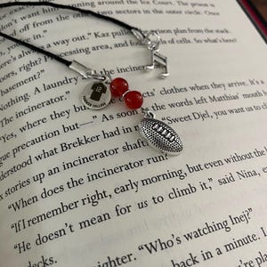 May include: A black cord bookmark with a football charm, red beads, and a silver charm with the number 12 and the words "Former College". A musical note charm is also attached.