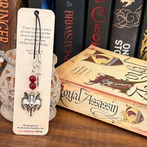 May include: A silver wolf head bookmark with red beads and a small sword charm. The bookmark is attached to a tan card with text from "Royal Assassin" by Robin Hobb. The card also includes the maker's name and social media handles.