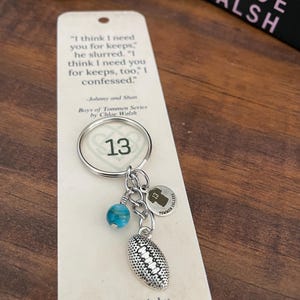 May include: A silver keychain with a football charm, a blue bead, and a charm with the number 13. The keychain is attached to a card with a quote from the book "Boys of Tommen Series" by Chloe Walsh.