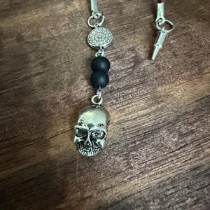 May include: A necklace featuring a silver-toned skull pendant, two matte black spherical beads, and a circular silver-toned charm with an intricate floral pattern. The pendant hangs from a thin black cord with a silver-toned metal bullet-shaped clasp.