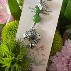 May include: A silver mushroom charm with a green bead and a small book charm hangs from a white cord. The charm is on a card with text and a mushroom illustration, surrounded by flowers.