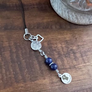 May include: A silver charm with a black cord, featuring a decorative design with two dark blue beads and a small round pendant. The pendant has an image of a person and the text "BIRTHDAY TRIBUTE".