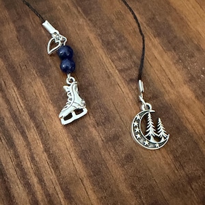 May include: Two bookmarks featuring black cords. One bookmark has a silver-toned ice skate charm, two dark blue spherical beads, and a small silver heart charm. The other bookmark features a silver-toned crescent moon charm with pine trees and stars.