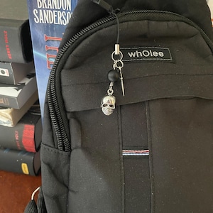 May include: Black sling bag with a skull charm. The bag has a zipper closure and a small pocket with the word "whoLee" printed on it. The charm is silver with a black bead and a small dagger.