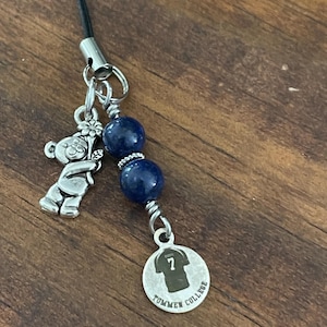 May include: A handmade charm featuring a silver teddy bear, blue beads, and a Tommen College pendant with a jersey number 7. The charm is attached to a black cord, perfect for a necklace or keychain.