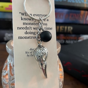 May include: Silver keychain with a raven skull charm, black bead, and a tag with text. The tag reads "When everyone knows you're a monster you needn't waste time doing monstrous". The keychain is next to a stack of books.