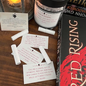 May include: A candlelit lantern, a jar labeled "Goblin Gospel," and several white paper scrolls with text are arranged on a wooden surface. A book titled "Red Rising" is visible. The items suggest a theme of fantasy or inspirational quotes.