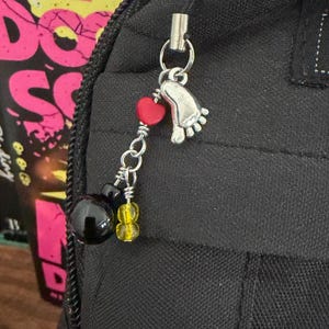 May include: A silver-toned metal charm featuring a foot-shaped pendant, a small red heart bead, a larger black spherical bead, and two smaller yellow translucent beads, all attached to a silver jump ring and a metal clasp on a black fabric surface.